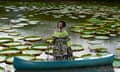 A woman stands in a canoe in a dress with African print. The canoe floats in a pond with scores of large lily pads.