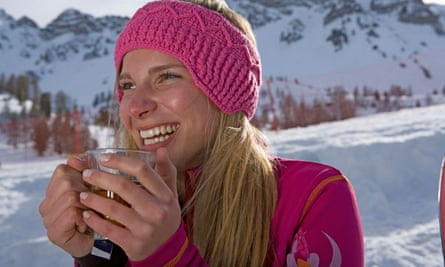 young woman with long blond hair, smiling, holding a drink and standing in front of a snowy ski slope. She is wearing a pink top and matching pink woolly headband