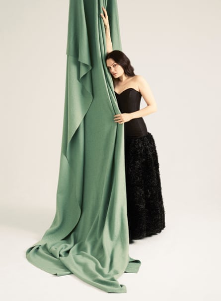 Jessie J wearing a full-length, strapless black dress standing against a long piece of pale-green material hanging from above her, with her right up held up straight against the material and her left hand gripping it in front of her body