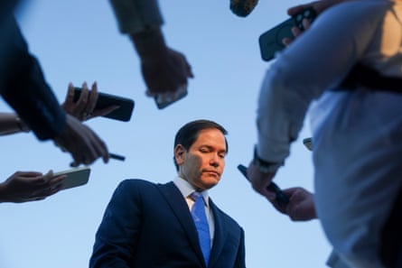 a man in a suit looks down as he is surround by people holding their phones toward him
