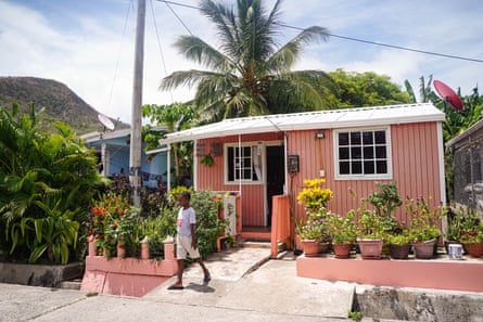 A small pink-painted single-storey house with a white corrugated roof. It has pots of flowers outside and a satellite dish. A young boy stands in front. There are palm trees behind it and a view to a steep hillside.