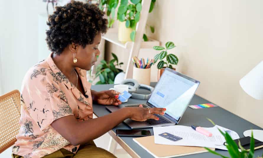 A woman at home in her home office sitting at a desk with a laptop online shopping. She is reading the details from her credit card as she makes an online purchase.