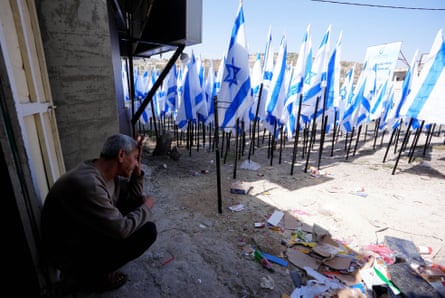 A man sits outside a house with dozens of Israeli flags stuck in the ground in front of him.