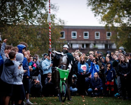 A person dressed in a knight costume rides atop a Lime bike in front of a crowd of young people in a park