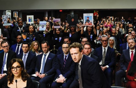 A solemn-looking young man in a suit sitting down while behind him protesters in the audience hold pictures of young people above their heads