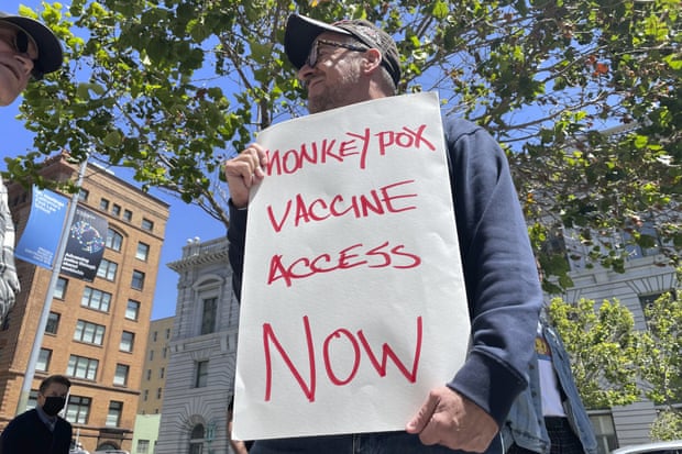 A man wearing a ballcap and glasses holds a sign reading ‘Monkeypox vaccine access now’.