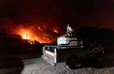 A man sits on top of a water tank with a generator mounted on the back of a truck, pumping water towards a blaze.