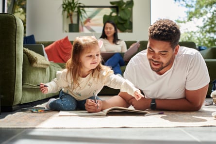 A father and daughter spend quality time together lying on the floor and writing in a notebook while the mother sits on the sofa in the background.
