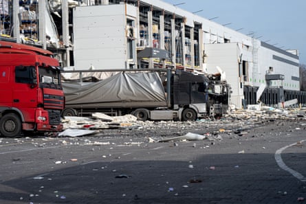 Damaged trucks in front of white industrial buildings, also damaged, with wreckage and rubble strewn across the carpark.