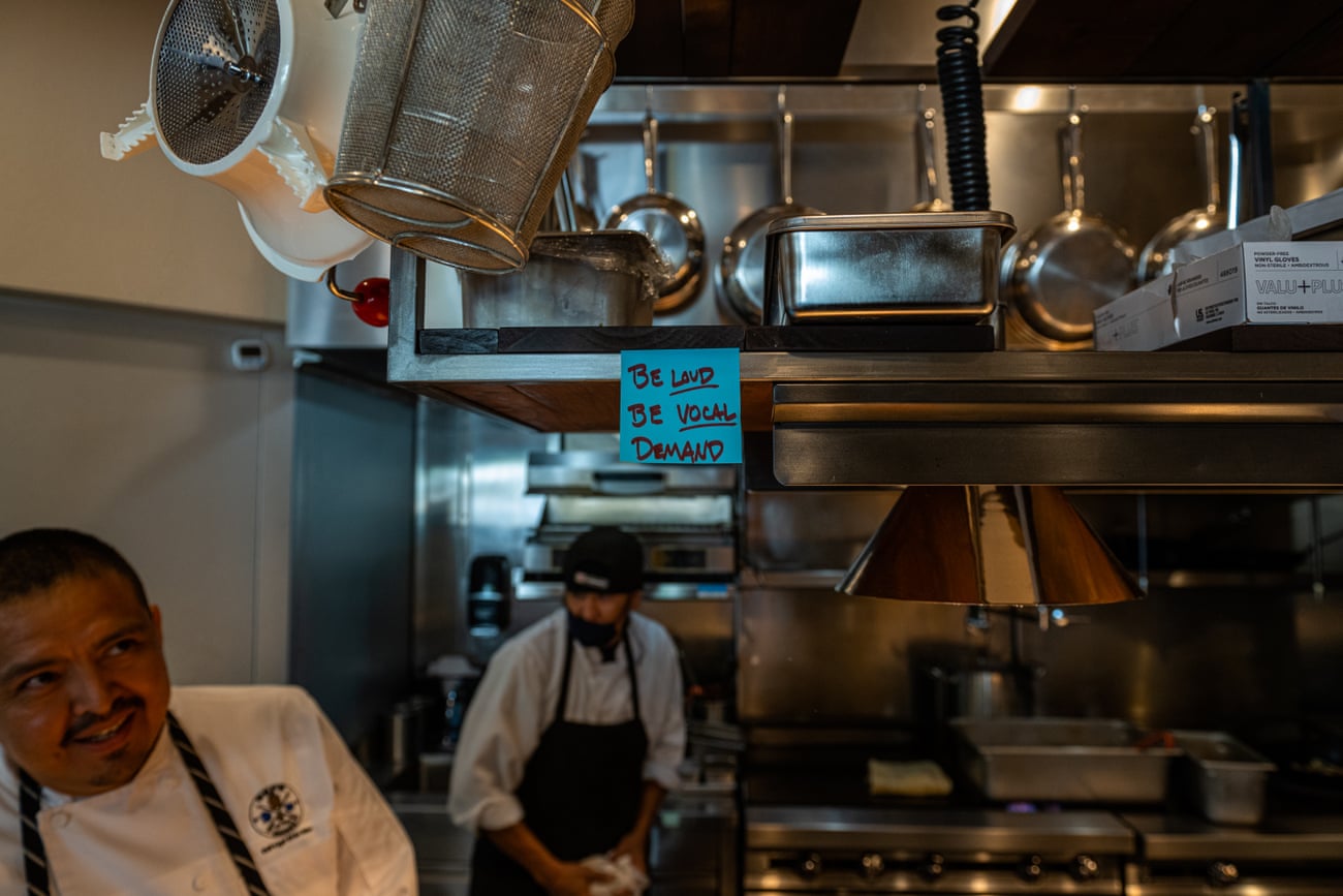 Two men work near stoves in a professional kitchen. A blue sticky note attached to an overhead shelf filled with shiny metal tools reads ‘Be loud, be vocal, demand!’