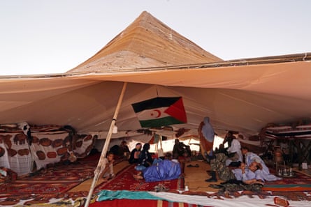 A group of people sit or recline under an open-sided Bedouin-style tent, with a striped flag hanging in the background
