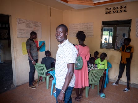 A young man smiles at the camera in a communal area with children and other adults around a table in the background