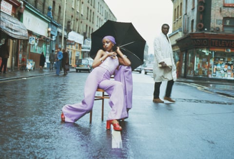 A woman in purple outfit sits on a chair in a wet Brixton Market street holding an umbrella, with a man walking past looking on