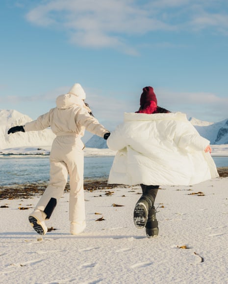 Two Cheerful Women Admiring Nordic Winter Walking at Scenic Beach on Lofoten IslandsRear view of two handsome happy female friends holding hands and running at majestic beach with snowcapped Mountain View under Arctic sun on Lofoten Islands, Northern Norway