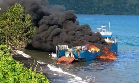 Smoke and flames come from a blue ship near a shoreline with mangroves