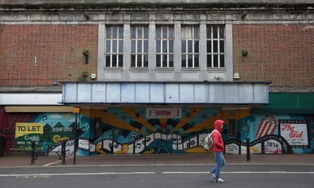 A person in a red hoodie and blue jeans walks past a boarded-up Odeon cinema. Graffiti-style artwork on the empty building includes a 'to let' sign