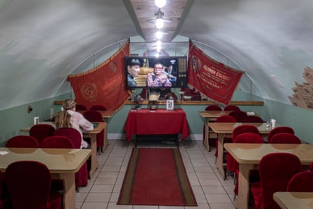 A person sits alone at a table in a restaurant watching a television at the far end of the room