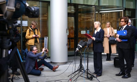 Sienna Miller, dressed in a suit, stands at a microphone outside a court building, holding a red folder, with camera crew and photographers in front of her