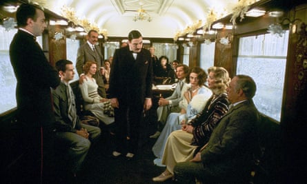 Finney dressed as Poirot with a florid moustache standing in the middle of a rail carriage with perimeter seating, surrounded by the other members of the cast in character.