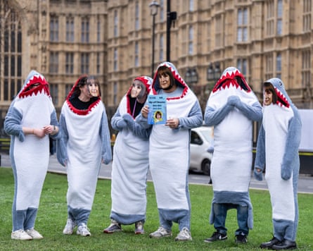 Six people in shark onesies stand on the grass in front of the Houses of Parliament