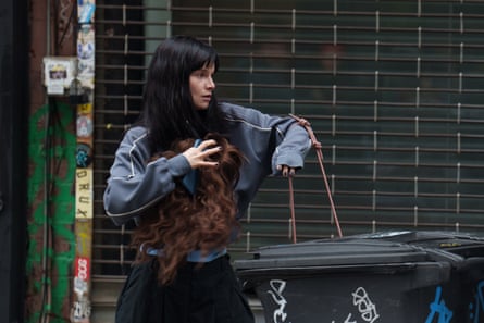 A young woman with long black hair and a fringe clutching a curly haired wig next to a rubbish bin
