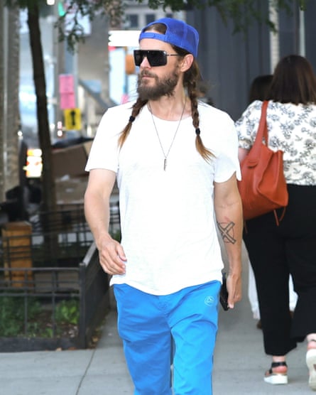 Leto walks in a New York Street. He is dressed casually in a white T-shirt and blue trousers, with a blue trucker cap worn back to front and and large retro sunglasses. His long brown hair is in two plaits and he has a beard.