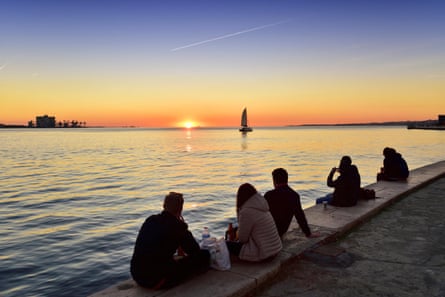 People sitting along the bank of a river at sunset with a yacht on the horizon