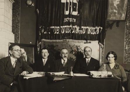 four men and a woman sit at a table under a banner with Yiddish writing on it