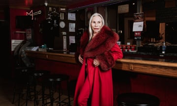 A blond woman  in a red velvet robe with a furry collar leans on a wooden bar