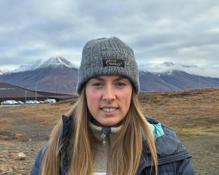 A young white woman stands outdoors in a beanie and jacket, with snow-capped mountains behind her