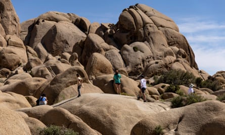 A line of people wearing T-shirts, shorts, and baseball caps walk across beige, bulbous rock formations against a blue sky.