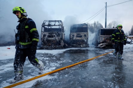 Two firefighters in front of burnt out trucks in a snowy urban setting