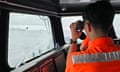 A man in orange hi-vis clothing peers through binoculars through the window of the vessel he is on at a boat in the distance