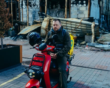 A man with a moustache sitting on a red vespa in front of a bombed-out building