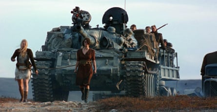 Two women wearing dresses walk in front of a tank and an army people carrier