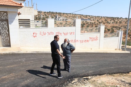 Two men stand in the road in front of a wall with writing sprayed in red
