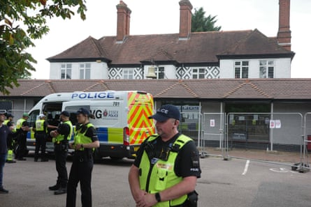 Police and a police van guard the hotel, a white-walled and tiled-roof building behind fencing