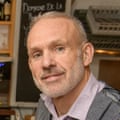 A portrait of Rob, with short grey hair, wearing a lilac shirt and a grey herringbone waistcoat
