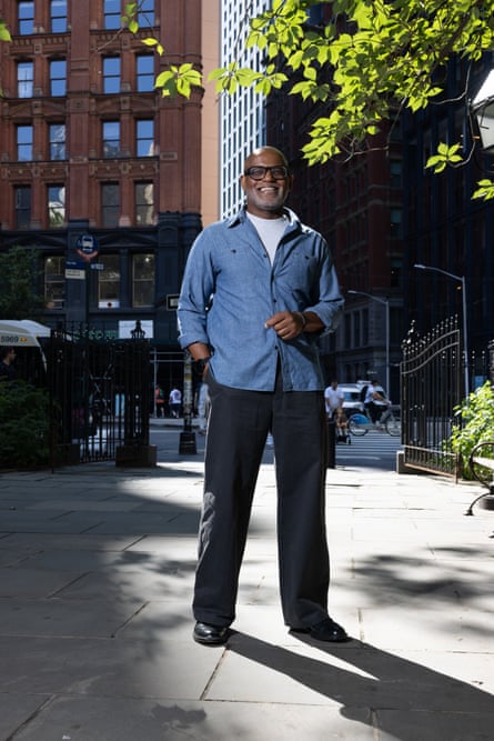 Gene Folkes standing in a park in front of office buildings, wearing a white T-shirt, a light-blue shirts, and black trousers and shoes, smiling at the camera