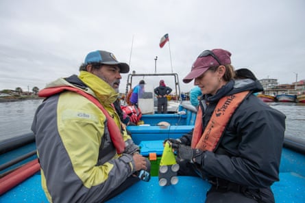 A man and a woman in waterproof jackets and life vests sit on a boat. The woman’s hands are on a small device with suckers and wires coming out of it on the bench between them