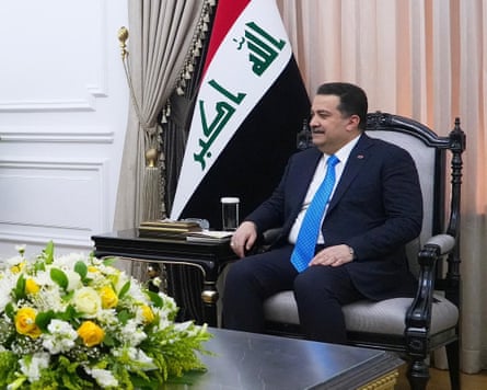 Mohammed Shia al-Sudani sits in a high wooden-framed chair in a meeting room; there is a large display of yellow and white flowers on the low table in front of him, and an Iraqi flag behind. He is in his 50s with short dark hair and moustache, and wears a dark suit and blue tie.