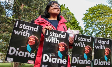 Diane Abbott stands behind a row of posters that feature her photo and the words ‘I stand with Diane’. A row of trees are in the background