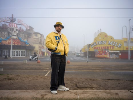 Young man with moustache and goatee wearing a hat and bright yellow jacket with Blackpool’s golden mile in the background.