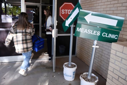 woman in plaid jacket and jeans walks into building past sign marked ‘measles clinic’ with an arrow