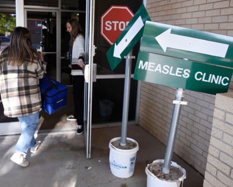 people walk into building beside green sign marked 'measles clinic' with an arrow