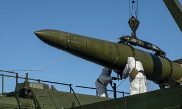 Two figures in white either side of a miltary-style green missile hanging against a blue sky