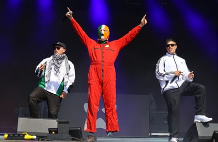 Kneecap perform at Glastonbury: the three are lined up on stage. Two wear sunglasses and white windcheater jackets, one with a keffiyeh scarf, with the central performer in a red boiler suit and a green, white and yellow balaclava.