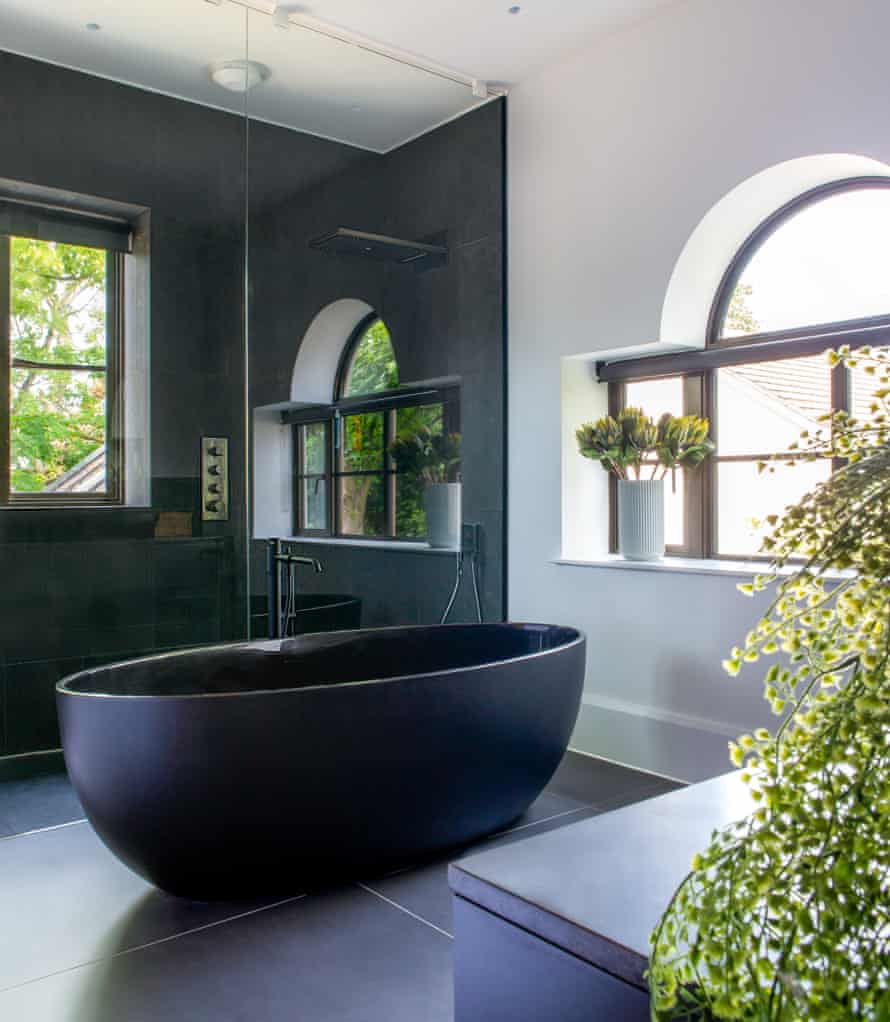 A deep black free-standing bath, in front of a glass-walled walk-in shower. There are two windows letting in light, and plants dotted around the surfaces.