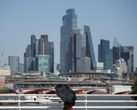 A person shields themself from the sun with an umbrella as they cross Waterloo Bridge with skyscrapers shimmering in heat haze beyond