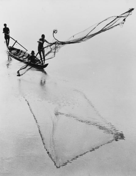 Three men are on a boat on a lake, one is throwing a net out to fish